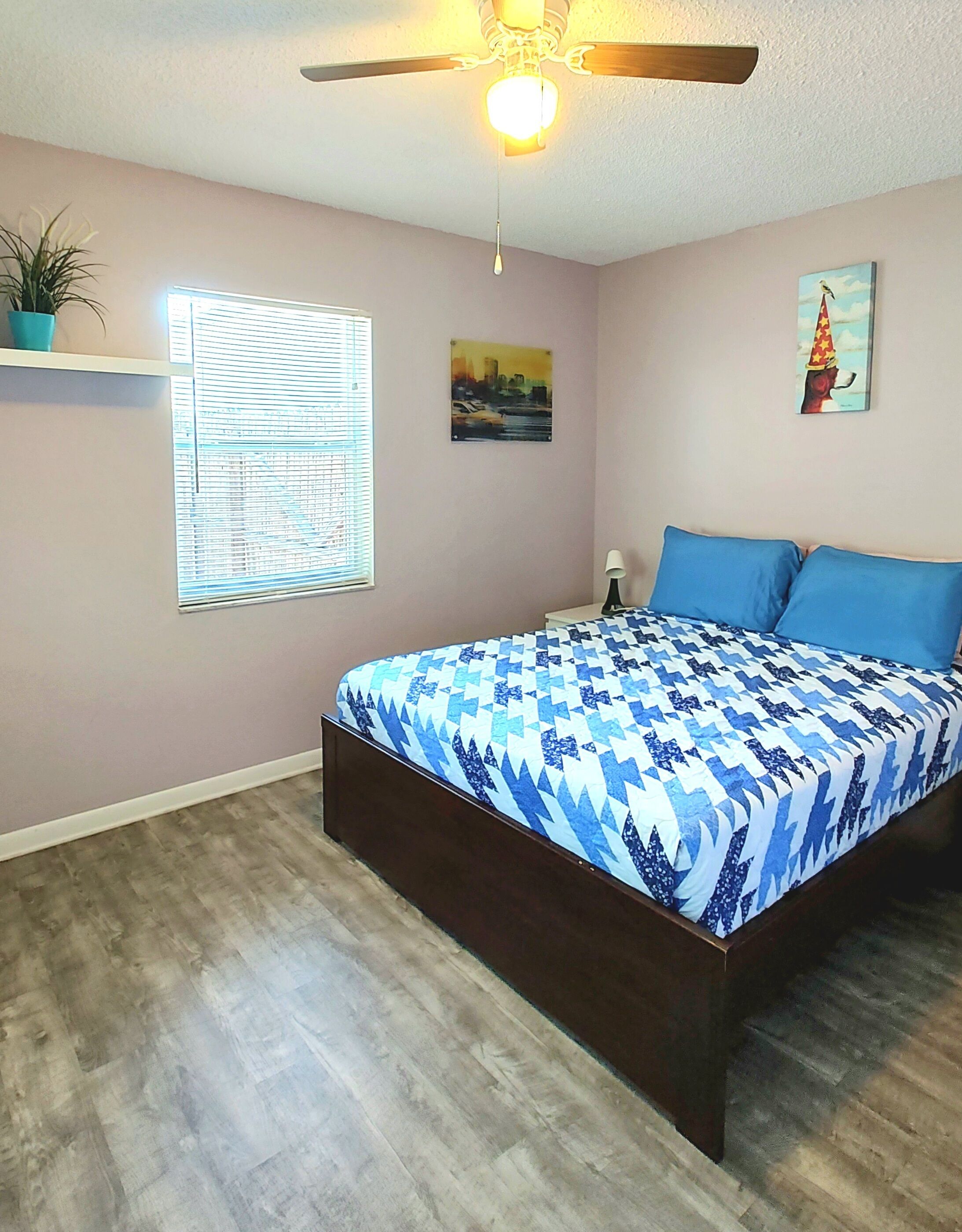 A bedroom with a bed, a ceiling fan, a window with blinds, a wall-mounted shelf with a plant, and a painting.