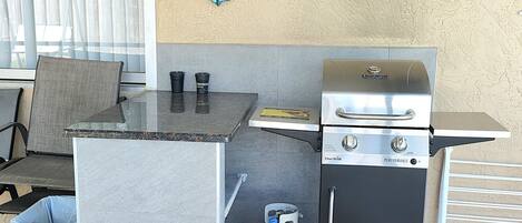 Un espace cuisine extérieur compact comprenant un barbecue, une table avec des chaises, une poubelle et une bouteille de propane.