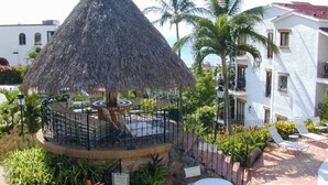 Un quiosco con techo de paja y zona de estar circular, rodeado por una cerca, palmeras y un edificio blanco con balcones.
