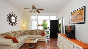 Una sala de estar con un sofá, televisión, ventilador de techo, un espejo con forma de sol y un cuadro.
