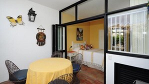 Una habitación con una mesa amarilla, sillas negras, una cama con cabecera blanca, un cuadro en la pared y una ventana con persianas blancas.