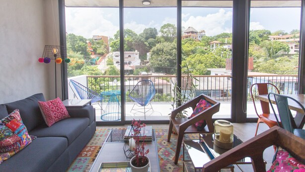 A modern living room with a grey sofa, colorful cushions, a coffee table with a plant, and a view of a cityscape through large windows.