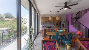 A modern kitchen with wooden cabinets, a dining area with colorful chairs, and a balcony with a view of the city.