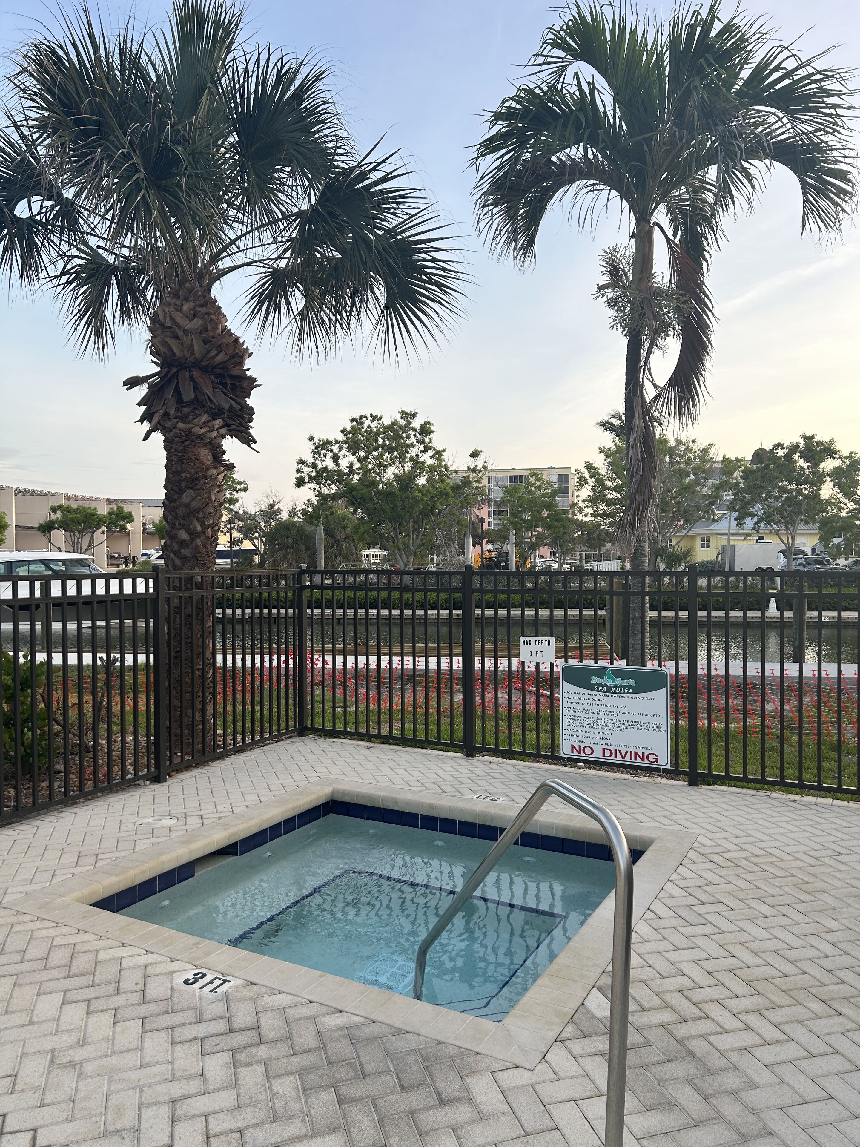 Zona de piscina con un letrero que indica una profundidad de 3 pies, rodeada de una valla, palmeras y vistas a un estacionamiento con coches y edificios.