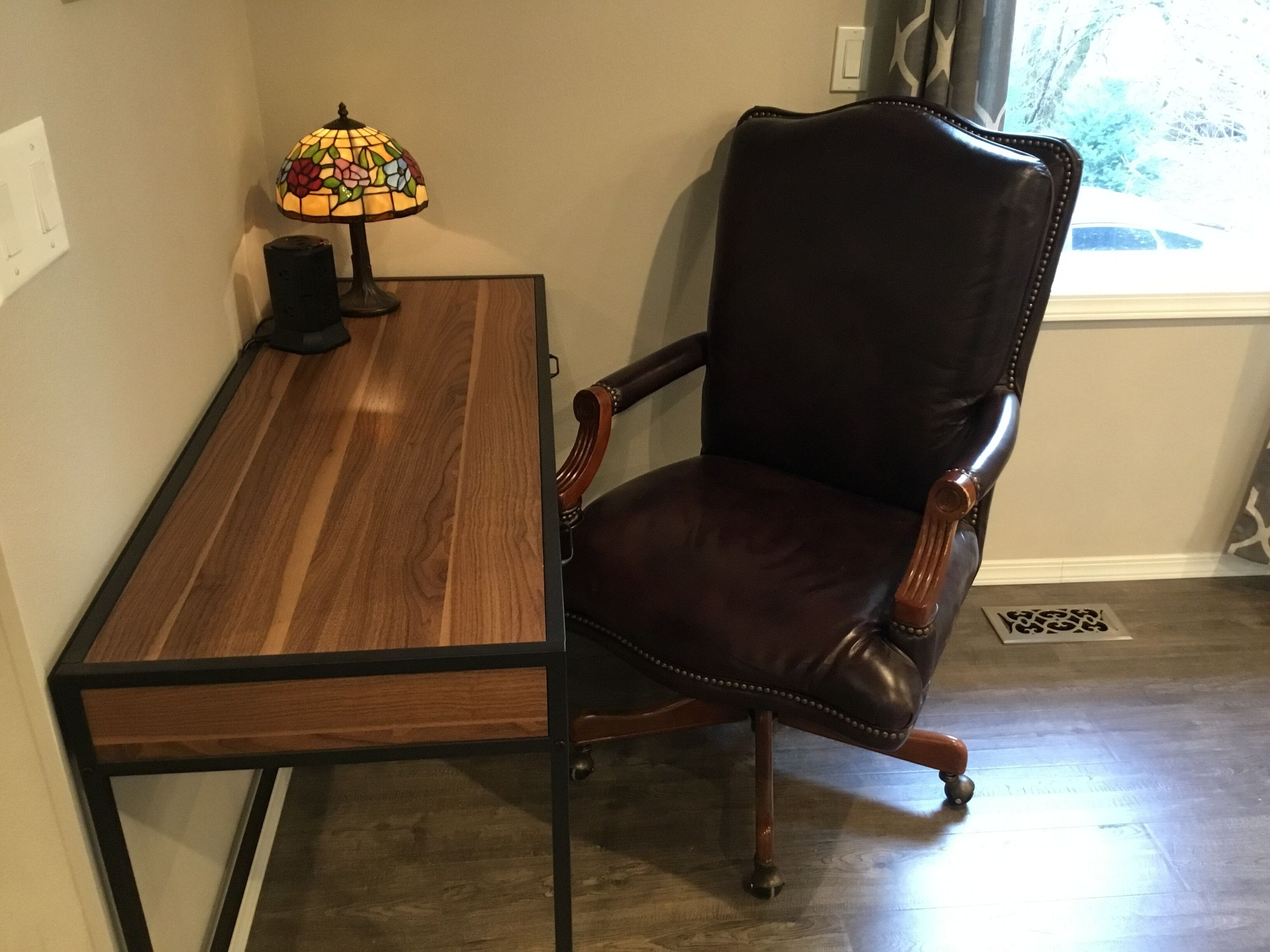 A wooden desk with a lamp, a leather chair, and a window with curtains.