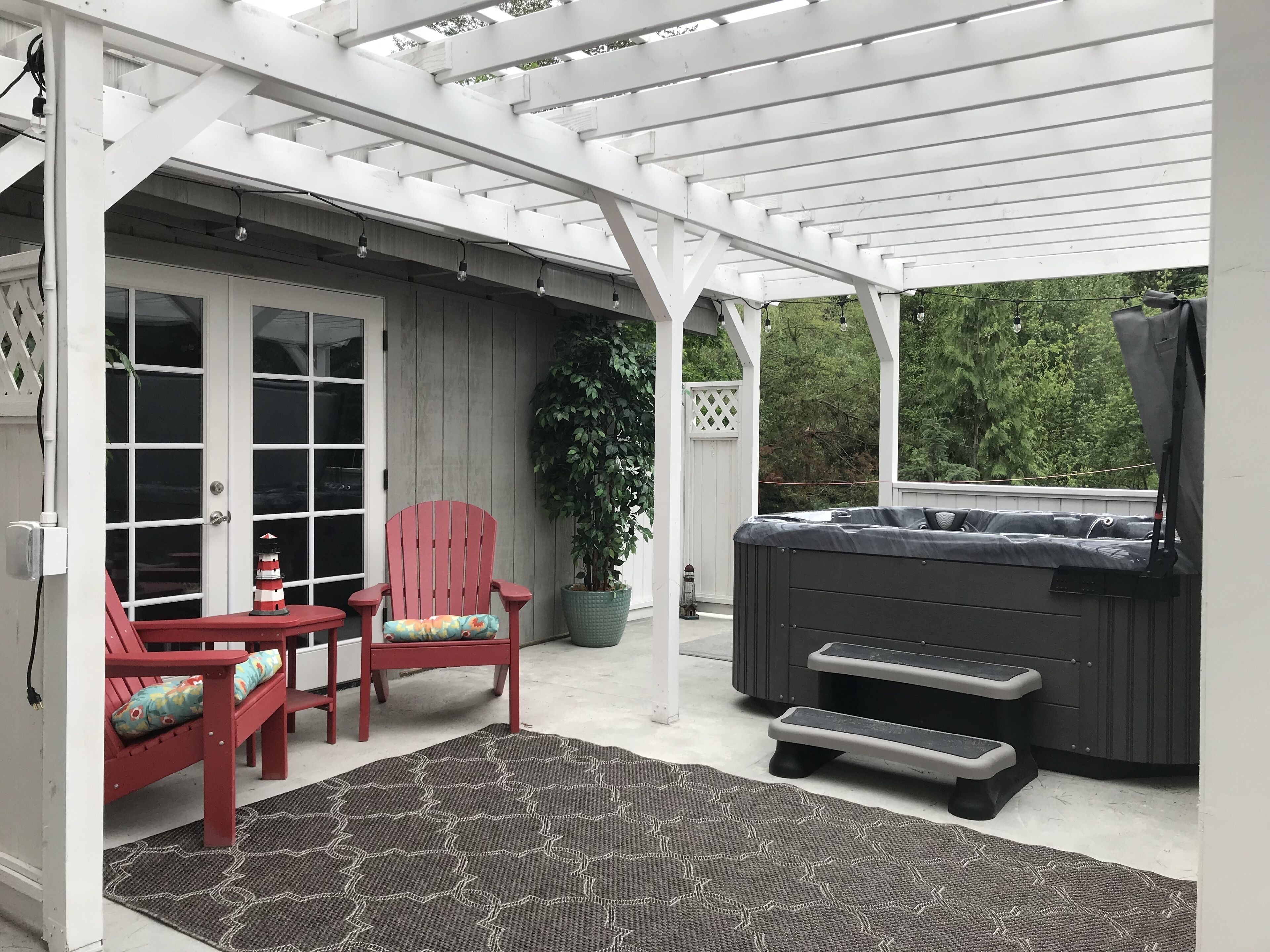 Un patio couvert comprenant un jacuzzi, deux fauteuils Adirondack rouges, une plante en pot et un tapis à motifs.