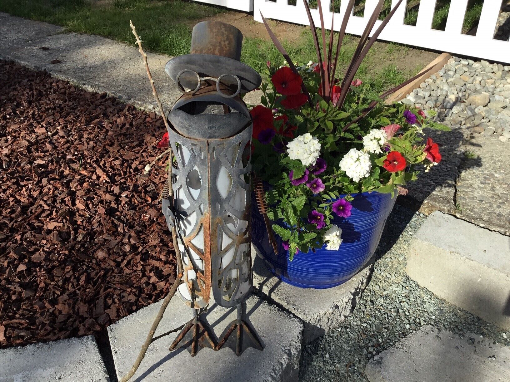 A metal lantern with a decorative top, placed on a concrete surface next to a flower pot with various flowers.