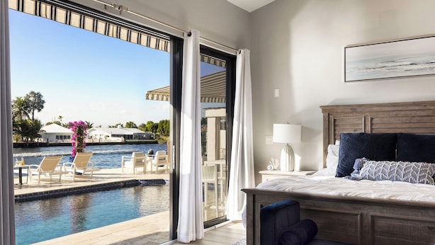 Ein Hotelzimmer mit einer großen Schiebeglaswand, die zu einem Poolbereich führt, einem Bett mit hölzernem Kopfteil und Blick auf den Pool sowie die umliegenden Gebäude.