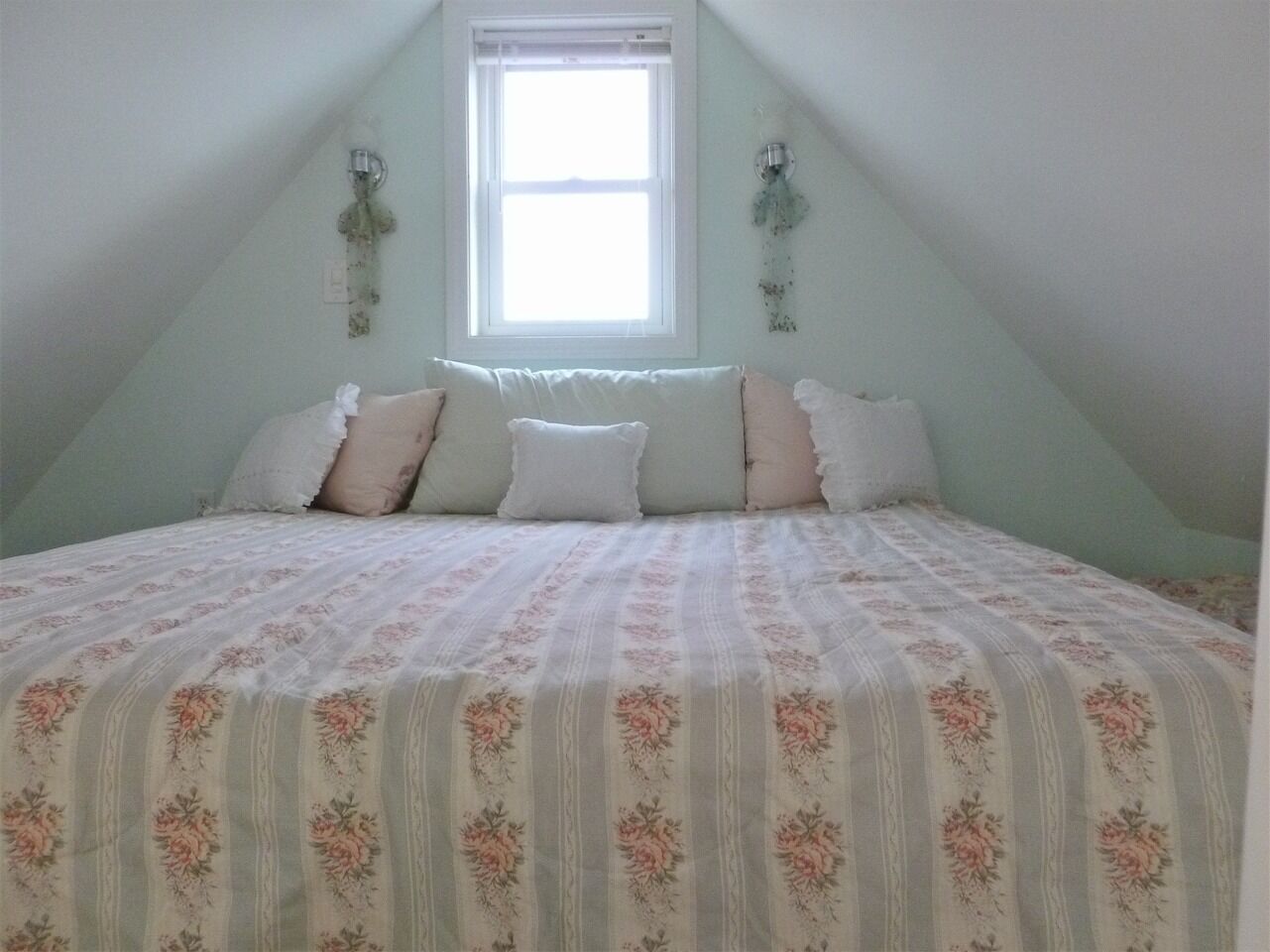 Un dormitorio con techo inclinado, una ventana y una cama con ropa de cama estampada con motivos florales.