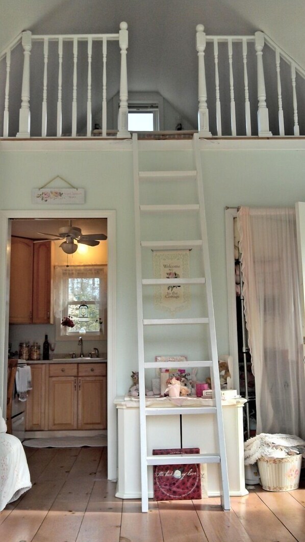 A loft bedroom with a white ladder leading to a lofted sleeping area. A kitchen is visible in the background. - Make Lasting Memories In This Adorable Vineyard Cottage! (Oak Bluffs)