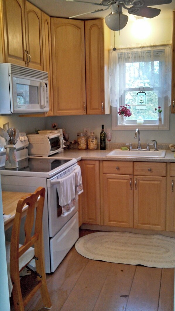 A kitchen with wooden cabinets, a microwave, a stove, a sink, and a window with a view of greenery. - Make Lasting Memories In This Adorable Vineyard Cottage! (Oak Bluffs)