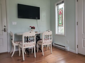 A dining area with a white table and two chairs, a flat-screen TV mounted on the wall, and a stained glass window. - Make Lasting Memories In This Adorable Vineyard Cottage! (Oak Bluffs)