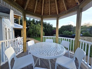 A covered outdoor area with a round table and white chairs, surrounded by a wooden deck and white railings. - Make Lasting Memories In This Adorable Vineyard Cottage! (Oak Bluffs)