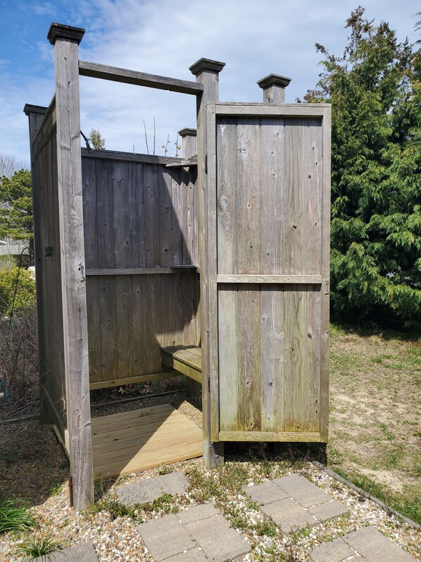 A wooden outdoor structure with a bench and a door, surrounded by gravel and greenery.