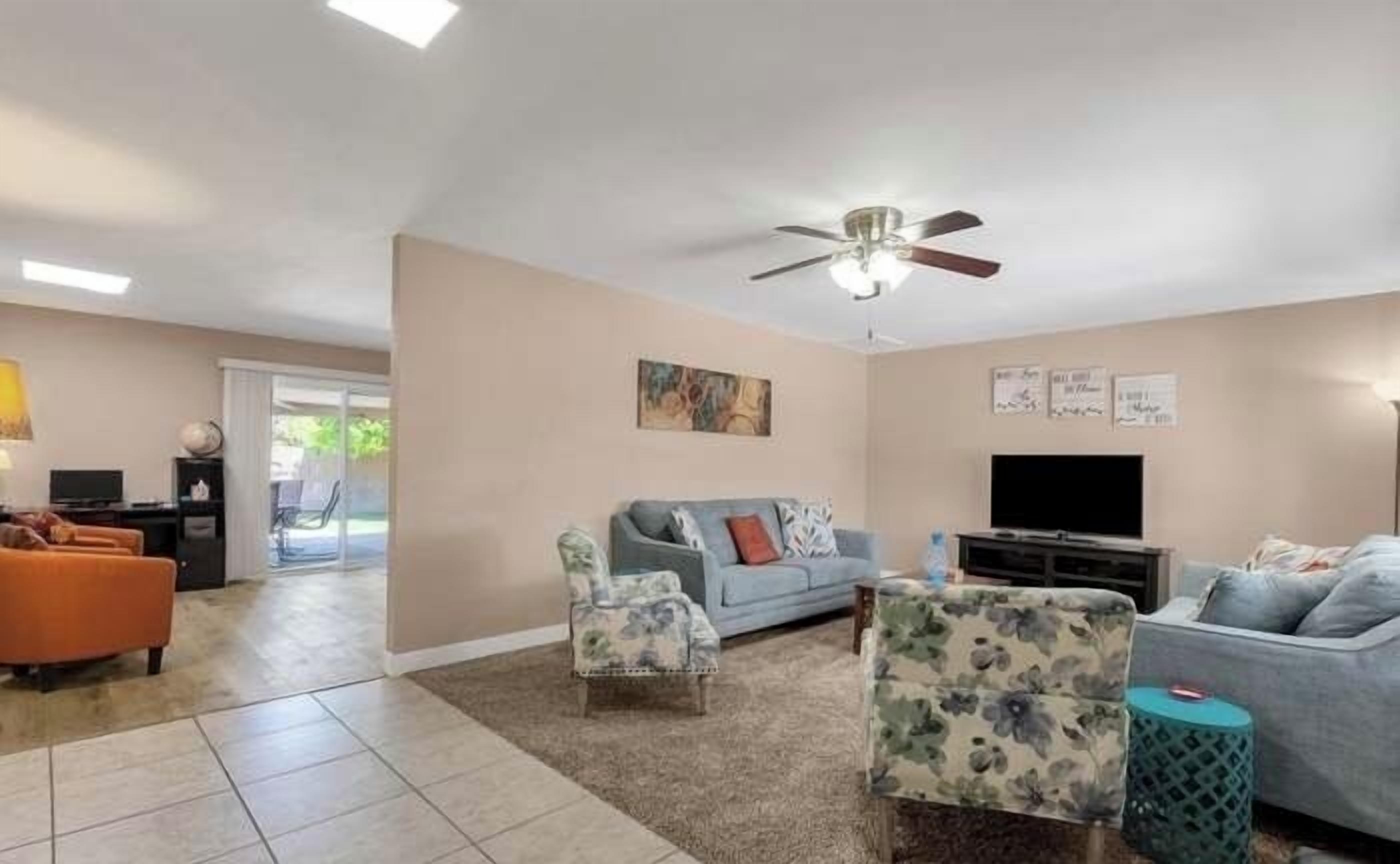 A spacious living room with a ceiling fan, a large flat-screen TV, and a comfortable seating area with a sofa and two armchairs.