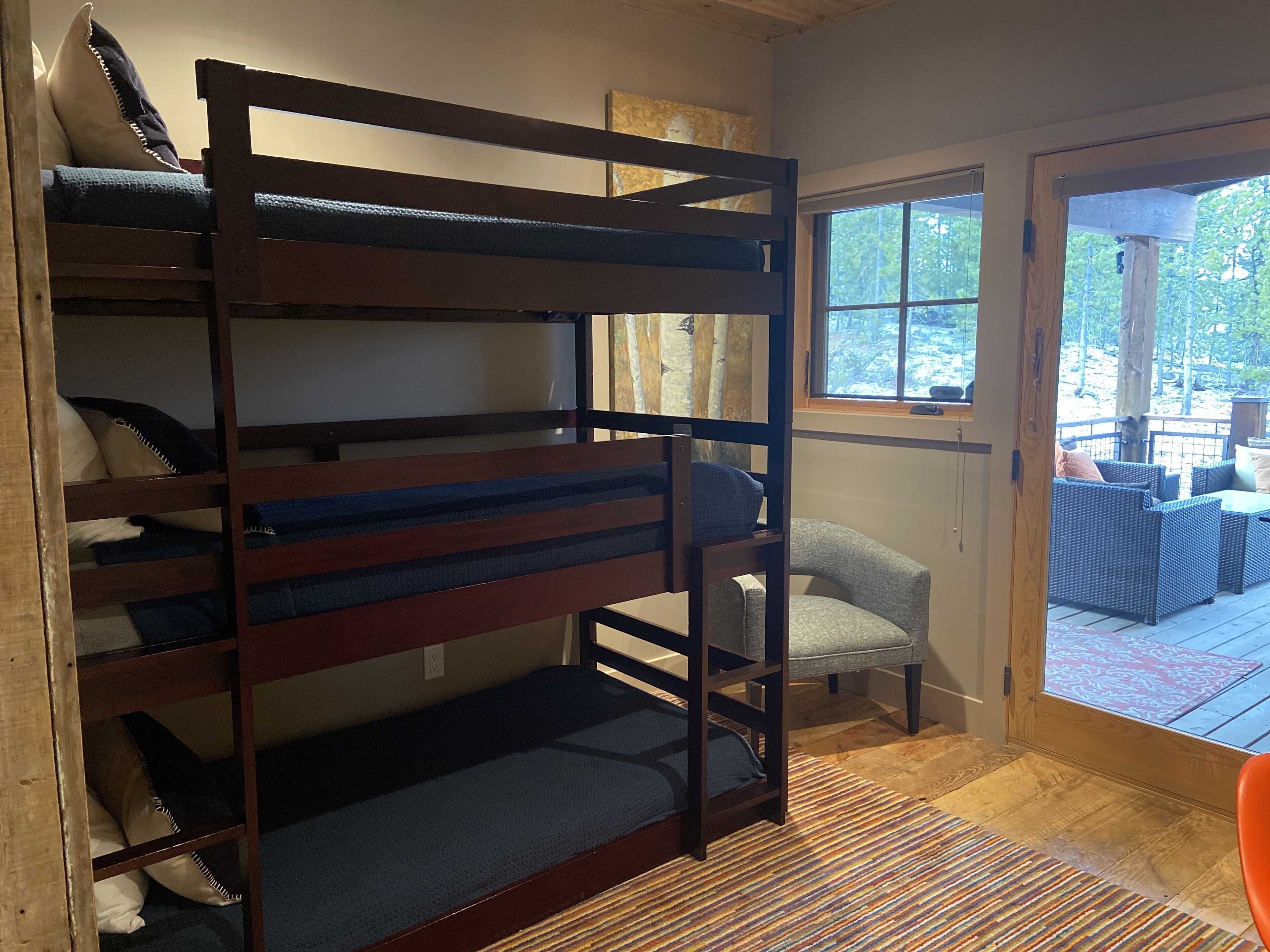 Una cama litera con ropa de cama azul, un sillón gris y una terraza de madera con mobiliario exterior.
