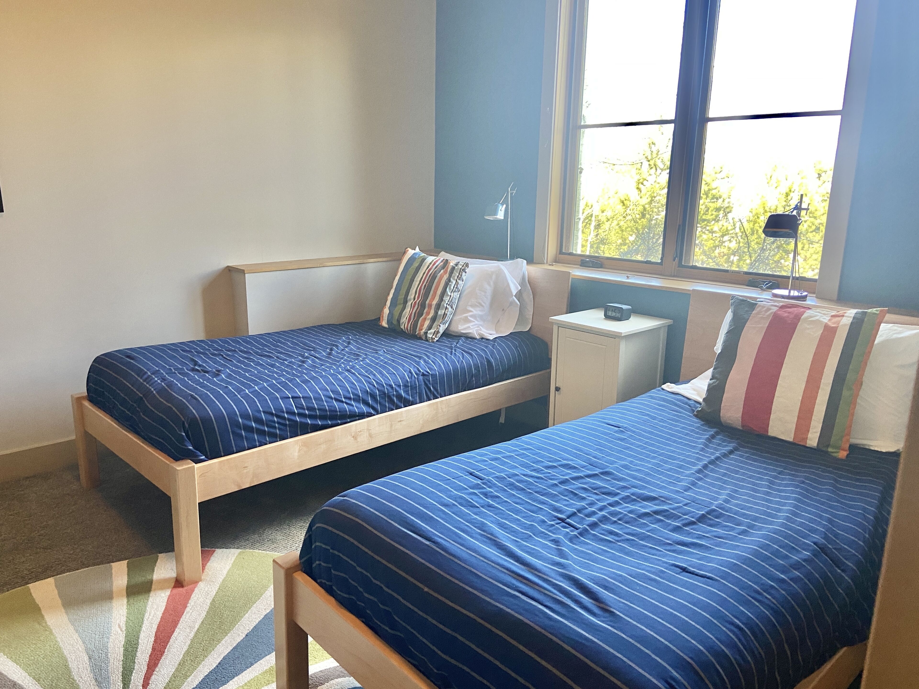 A bedroom with two single beds, a small bedside table with a lamp, a window with a view of trees, and a striped rug on the floor.