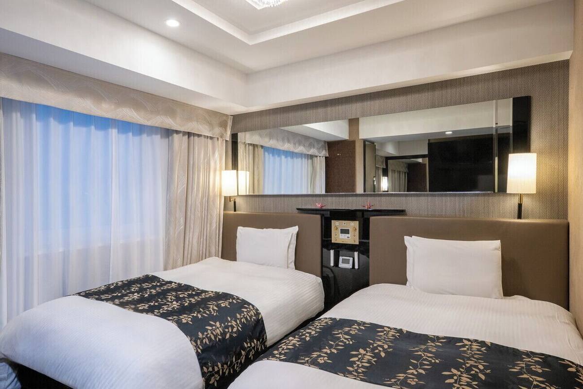 Habitación de hotel con dos camas, un espejo grande, un escritorio con panel de control y vistas al exterior.