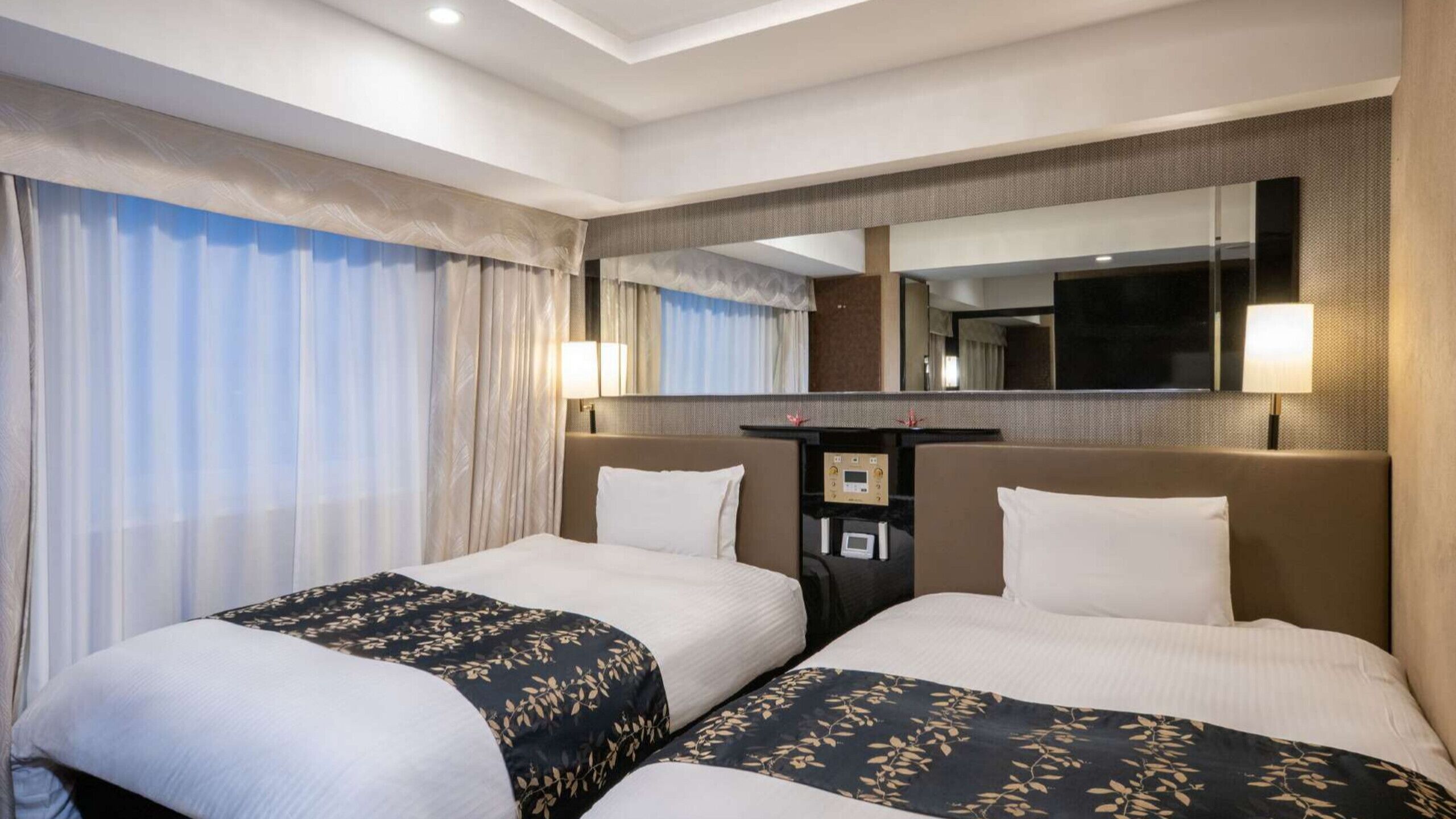 A hotel room with two beds, a large mirror, a desk with a lamp, and a window with curtains.