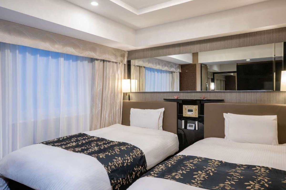 Habitación de hotel con dos camas, un espejo grande, un escritorio con lámpara y una ventana con cortinas.