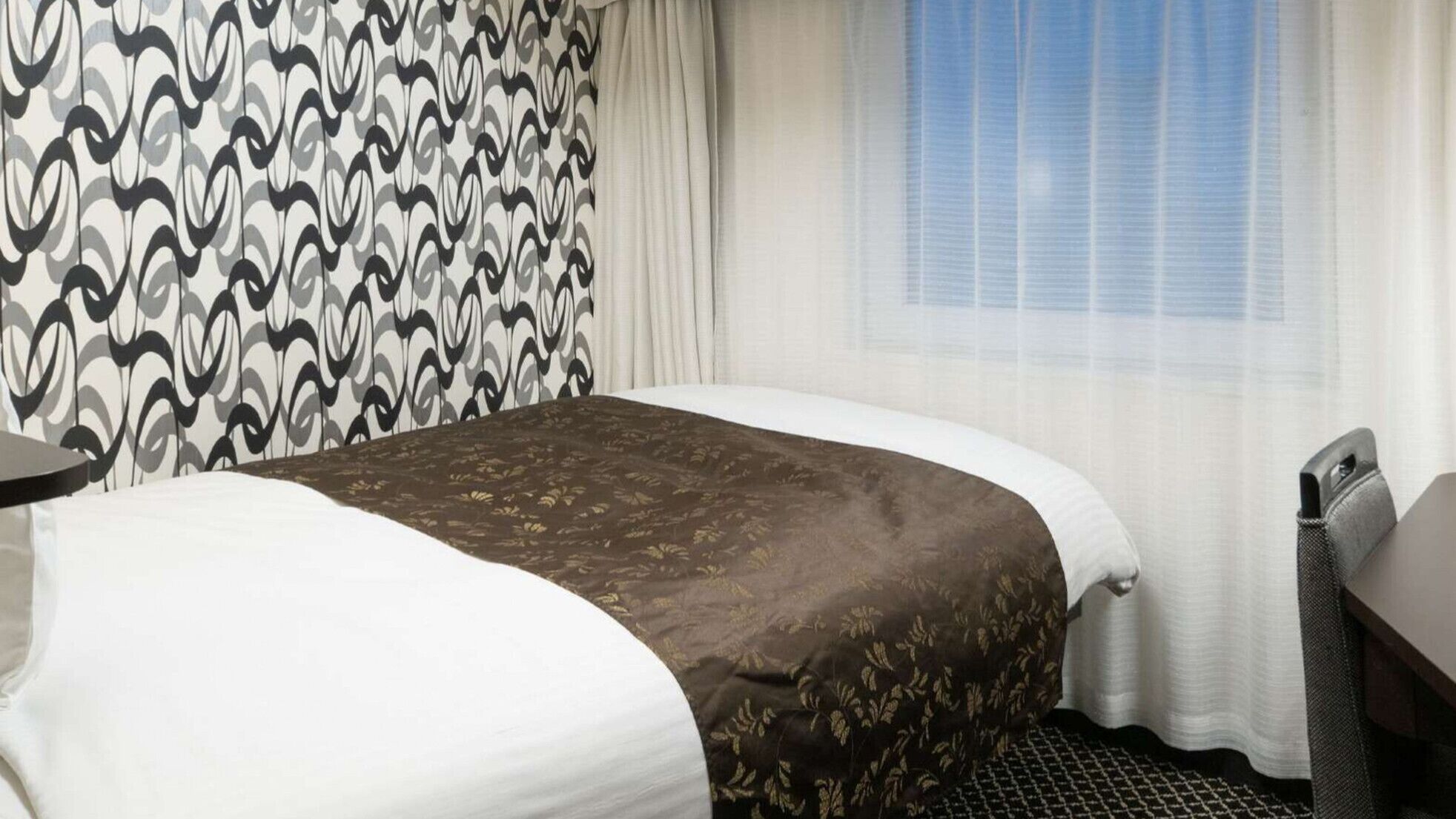 A hotel room with a bed, a bedside table, a chair, and a window with curtains.