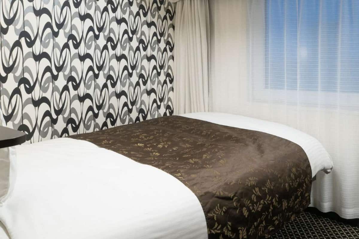 Una habitación de hotel con cama, mesita de noche, silla y ventana con cortinas.