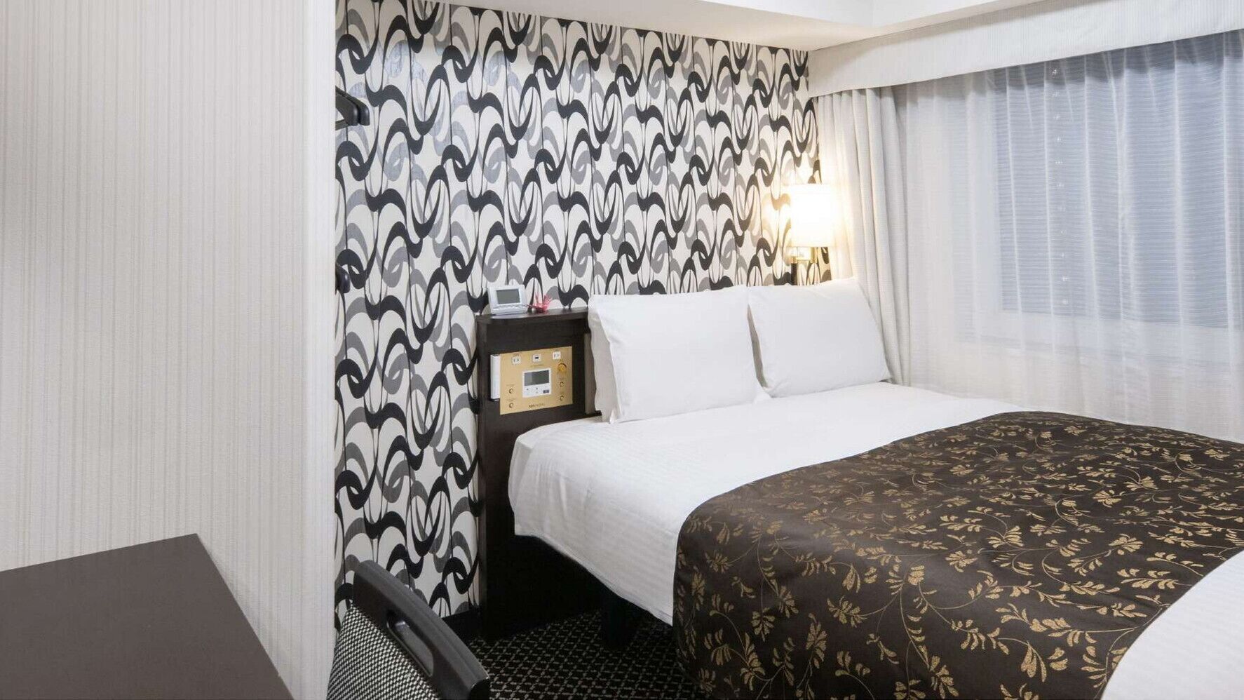A hotel room with a bed, a desk, a chair, a nightstand with a lamp, and a patterned wall.