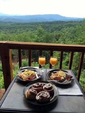 A table with breakfast plates, two glasses of orange juice, and cinnamon rolls on a balcony with a forest view. - Avail 10/2-6, 25-30, 11/8-30 Knight of My Dreams, Secluded, Amazing View,1 level (Sevierville)