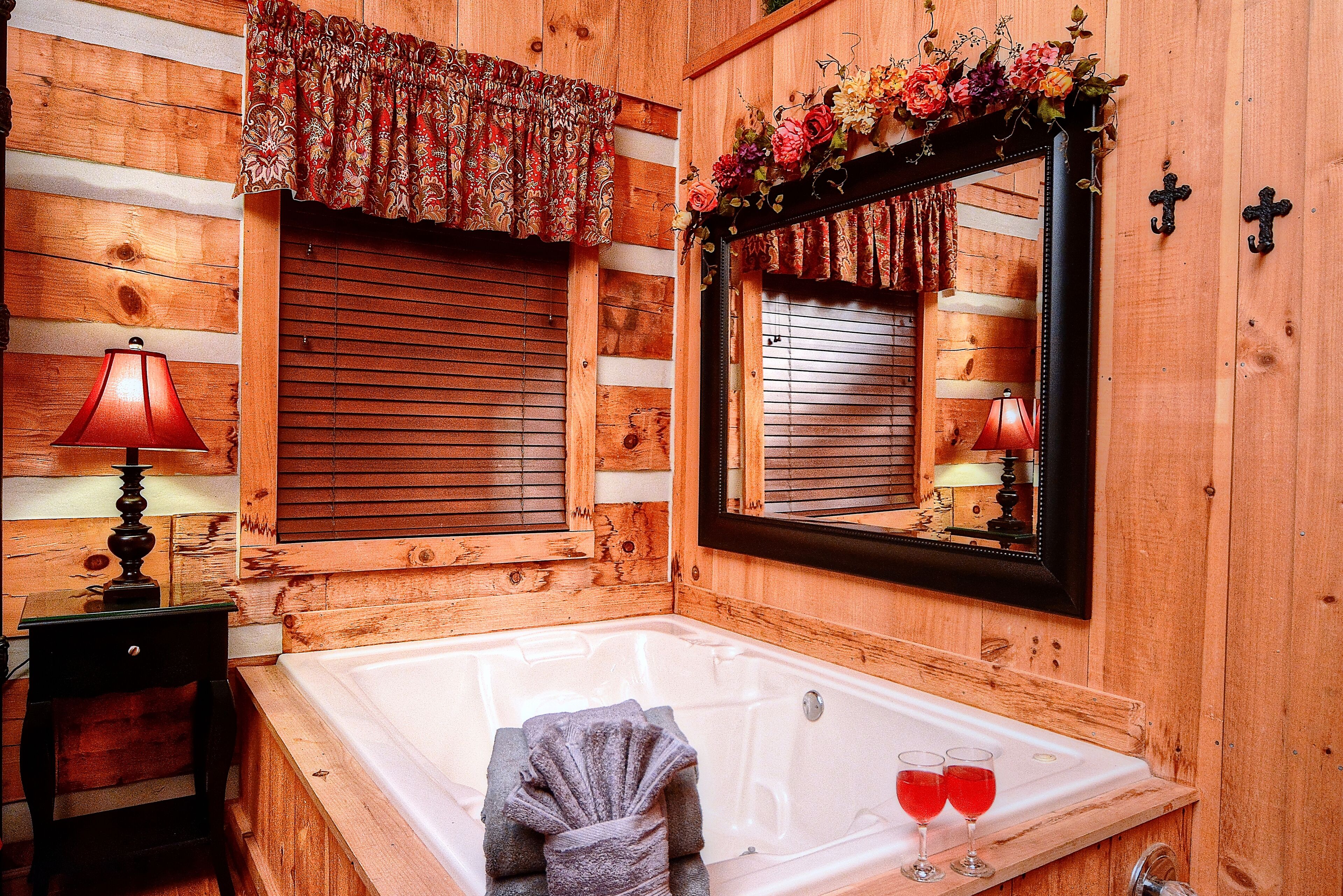 A wooden cabin bathroom with a bathtub, a mirror, a window with shutters, and a lamp.
