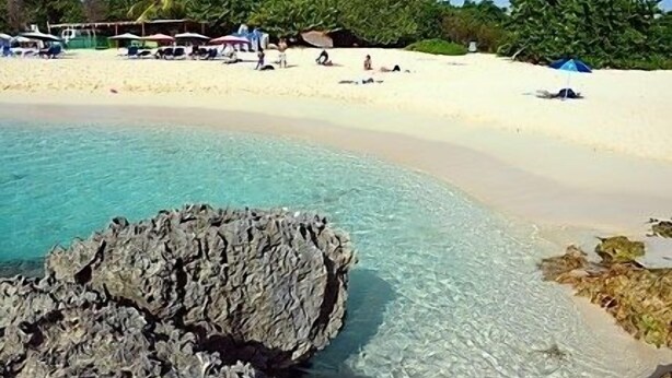 Een strand met helder turquoise water, wit zand en een rotsformatie op de voorgrond. Er staan enkele palmbomen en in de verte zijn mensen te zien.