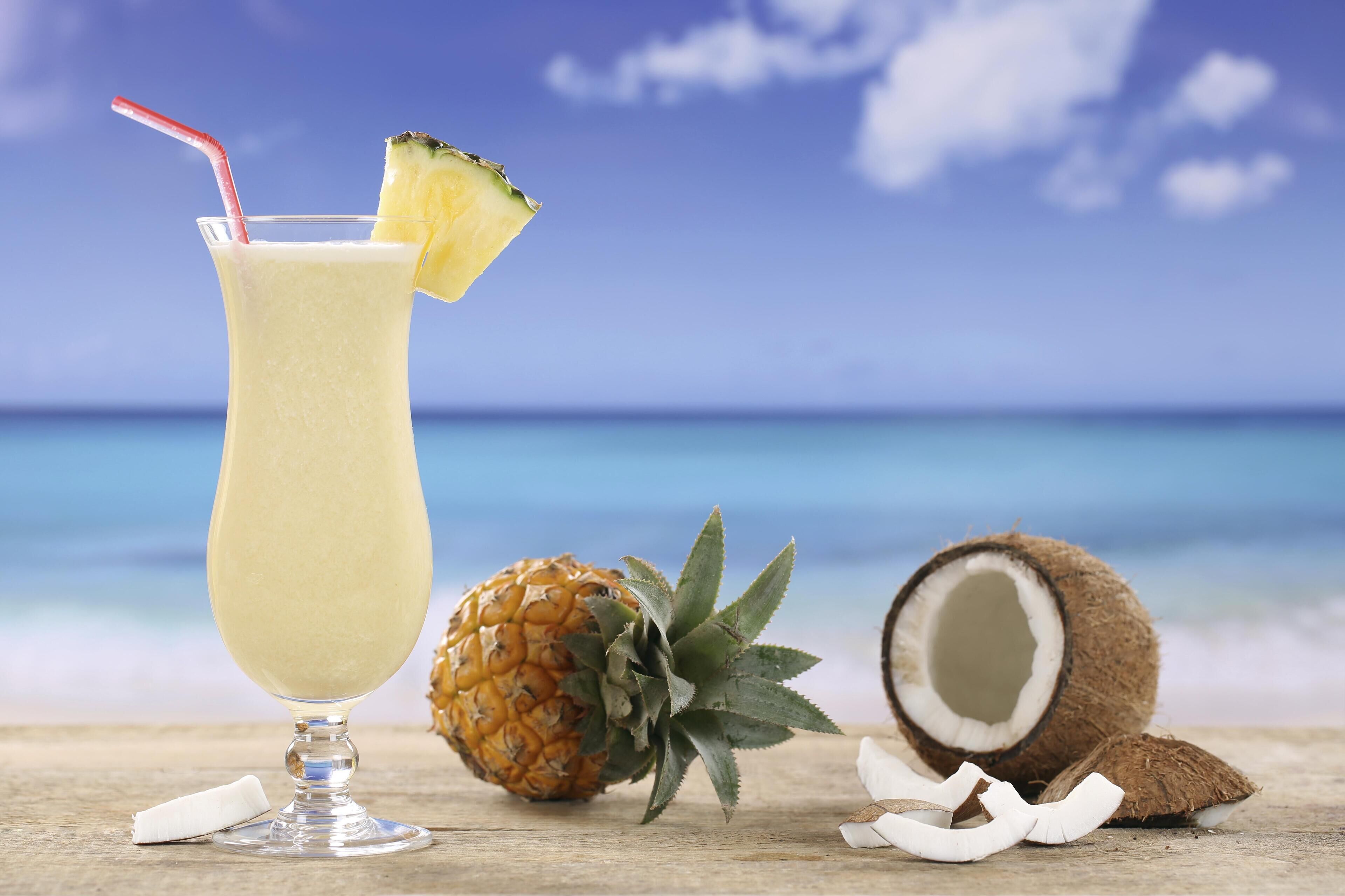 A tropical drink with a straw, a pineapple slice, and a coconut on a wooden surface with a beach and ocean in the background.