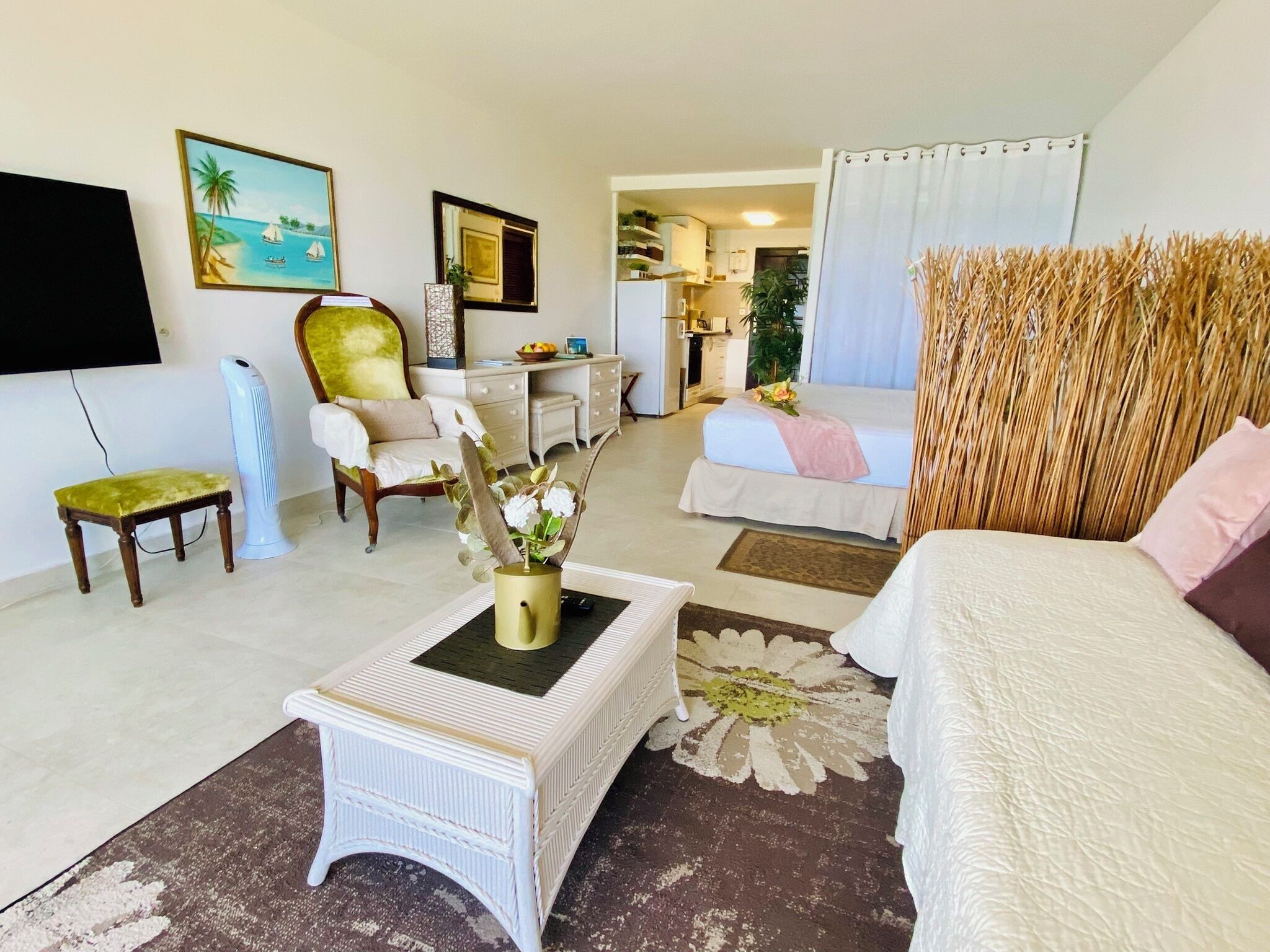 Ein helles, modernes Wohnzimmer mit Sofa, Couchtisch mit Vase, Wand-Fernseher, Essbereich mit Tisch und Stühlen sowie ein Schlafzimmer mit Bett und Vorhängen.
