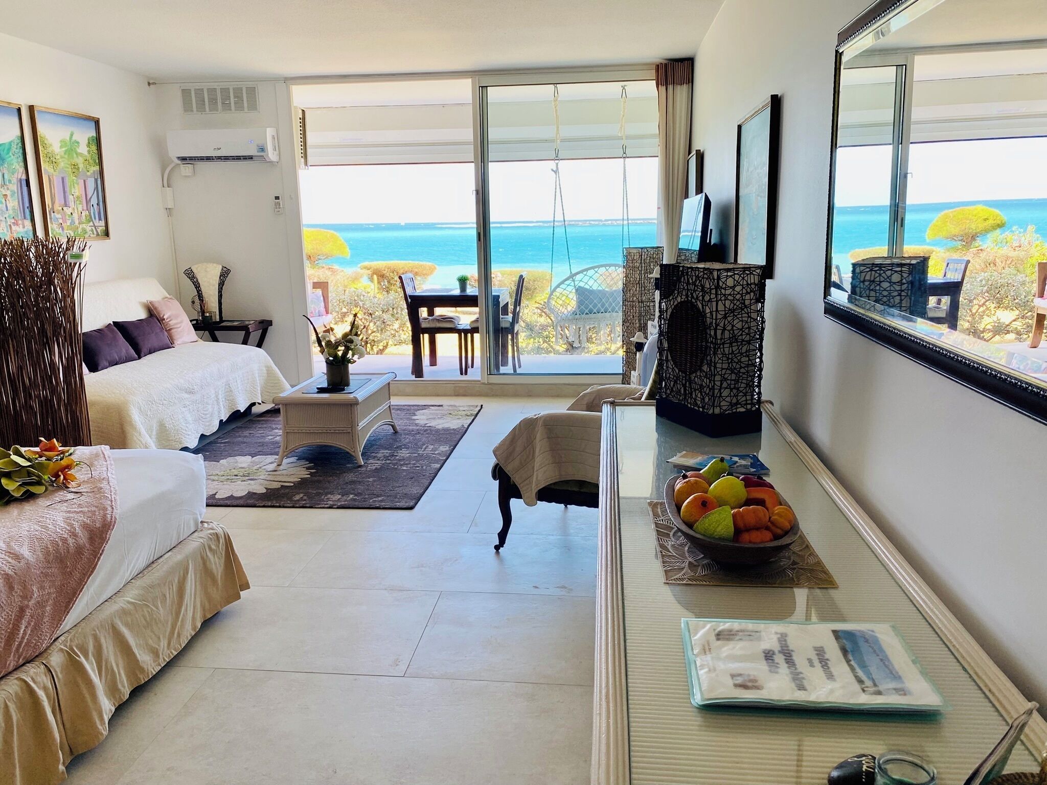 A hotel room with a bed, a desk with a bowl of fruit, a chair, a small table, and a view of the ocean through large windows.
