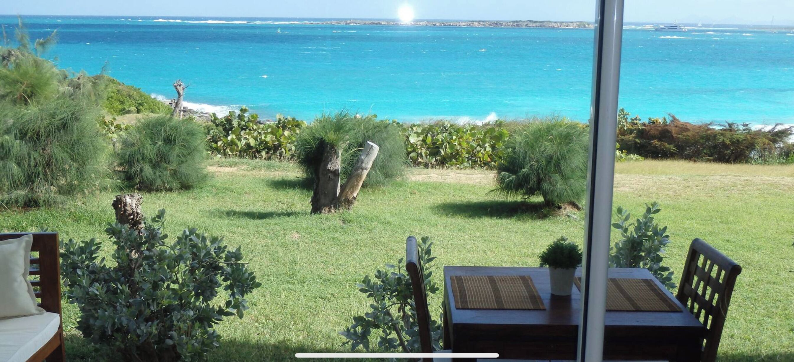 A patio with a table and chairs, overlooking a coastal view with the ocean and a grassy area with plants.