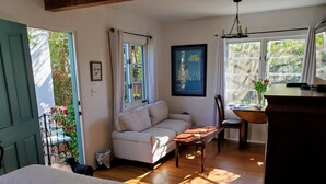 A cozy room with a bed, a sofa, a dining table, and a chair. There are windows with curtains, a framed picture on the wall, and a small trash bin. - Charming Guesthouse Studio To  Elegant 1920s Spanish Home (Coronado)