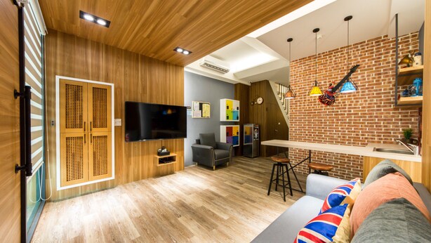 A modern living room with a wooden floor, a grey sofa, a TV, and a kitchen area with a brick wall and hanging lights. - Pickup Light (Tainan)