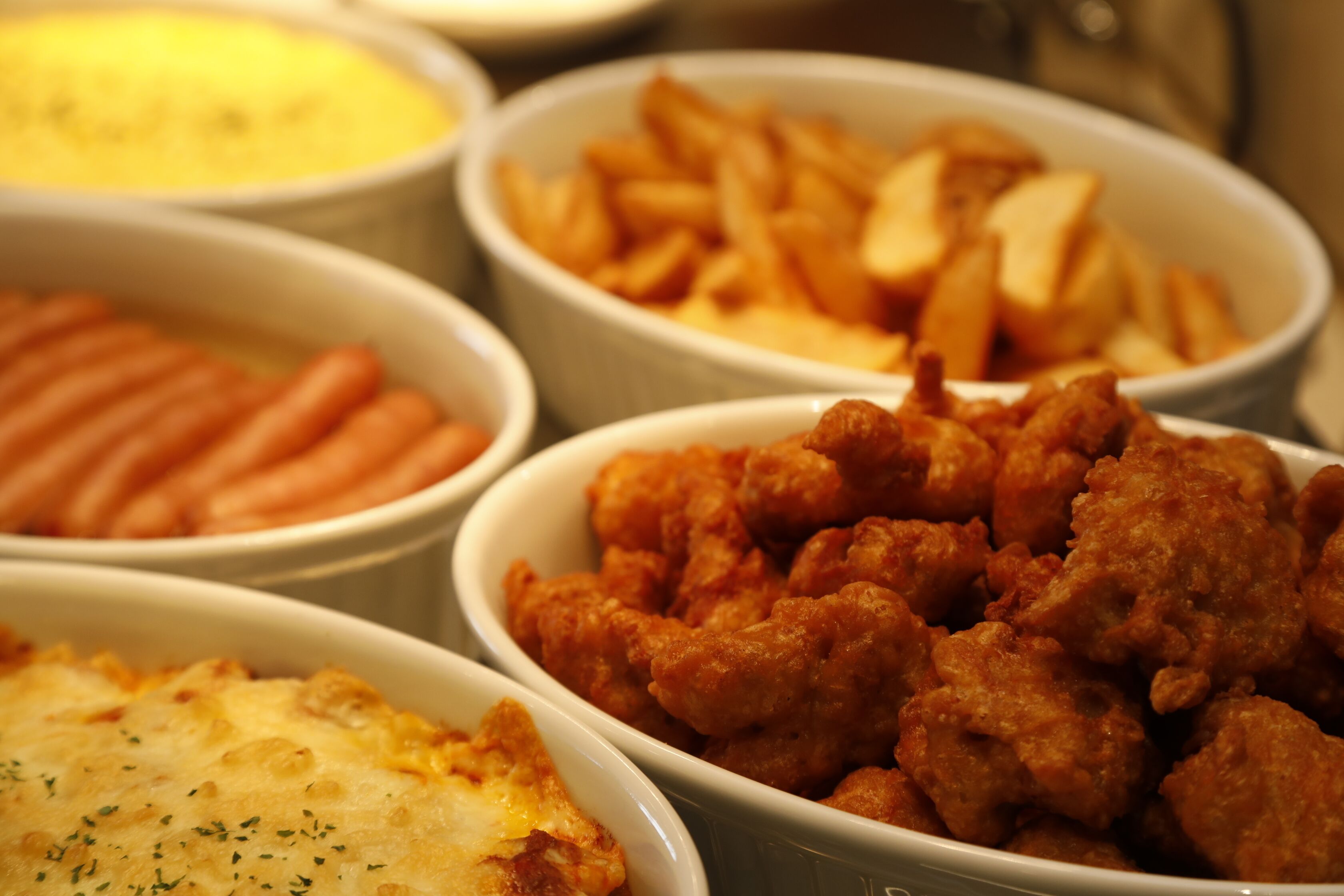 A variety of food items in bowls, including what appears to be French fries, a dish with sausages, and a fried chicken dish.