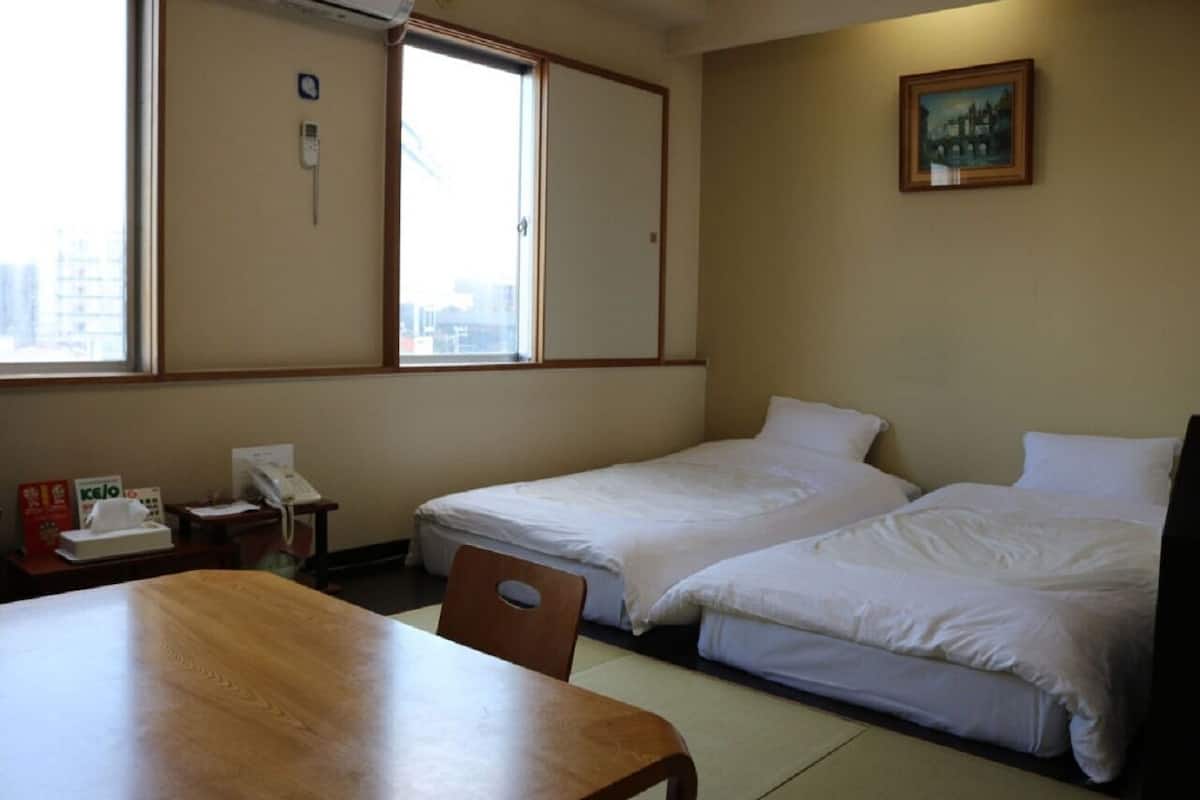 Quarto de hotel com duas camas de solteiro, uma mesa de madeira, uma cadeira, uma escrivaninha com telefone e uma janela com vista para edifícios.