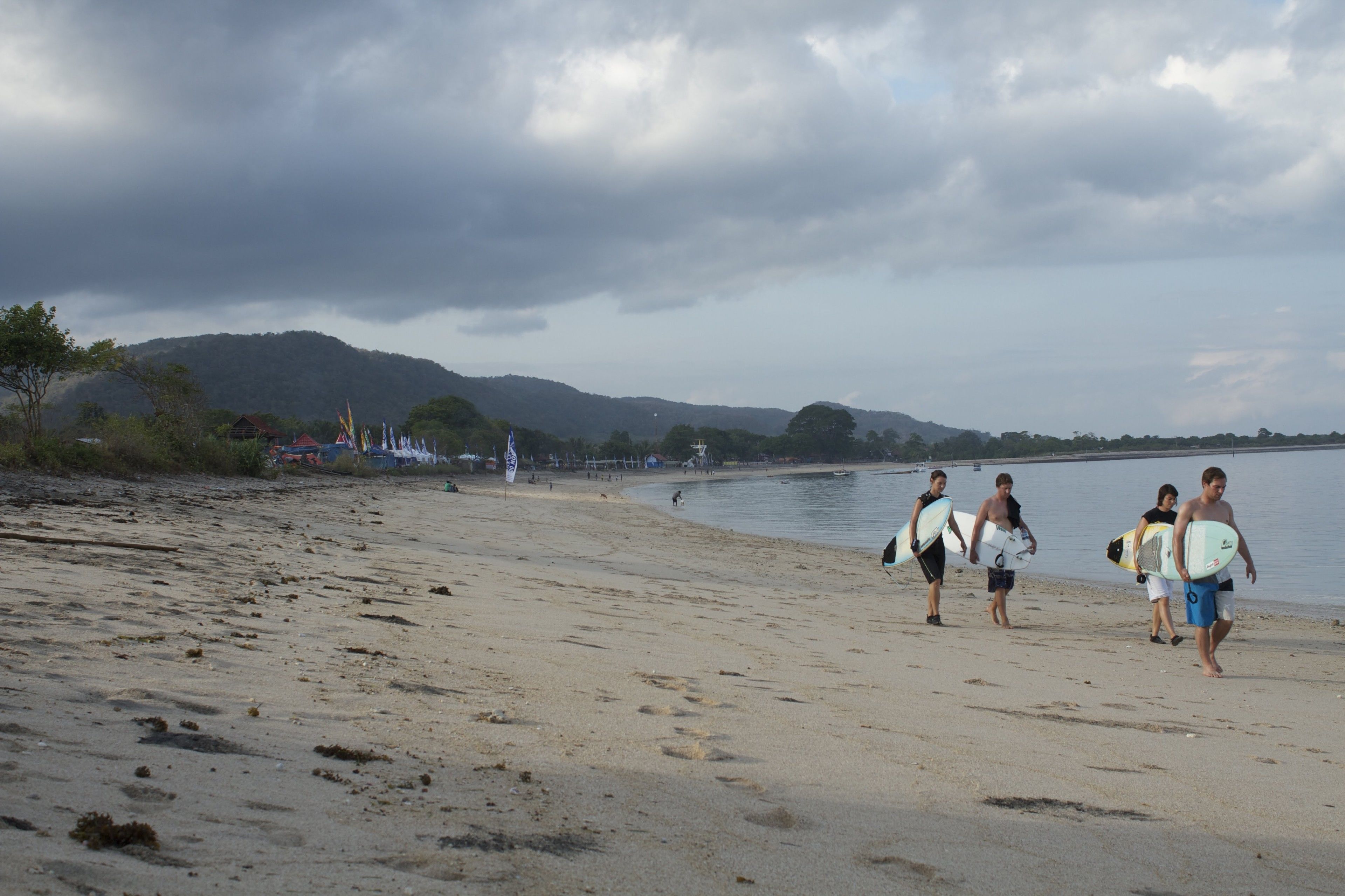 A beach with people walking and carrying surfboards, a calm sea, and distant hills.