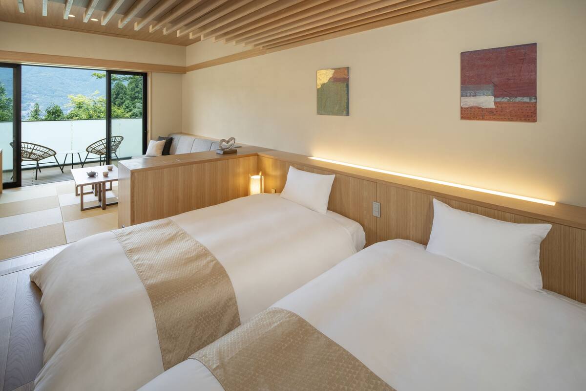 Une chambre d’hôtel avec deux lits, une tête de lit en bois, une petite table avec des chaises et une vue sur l’extérieur, accessible par une porte coulissante.