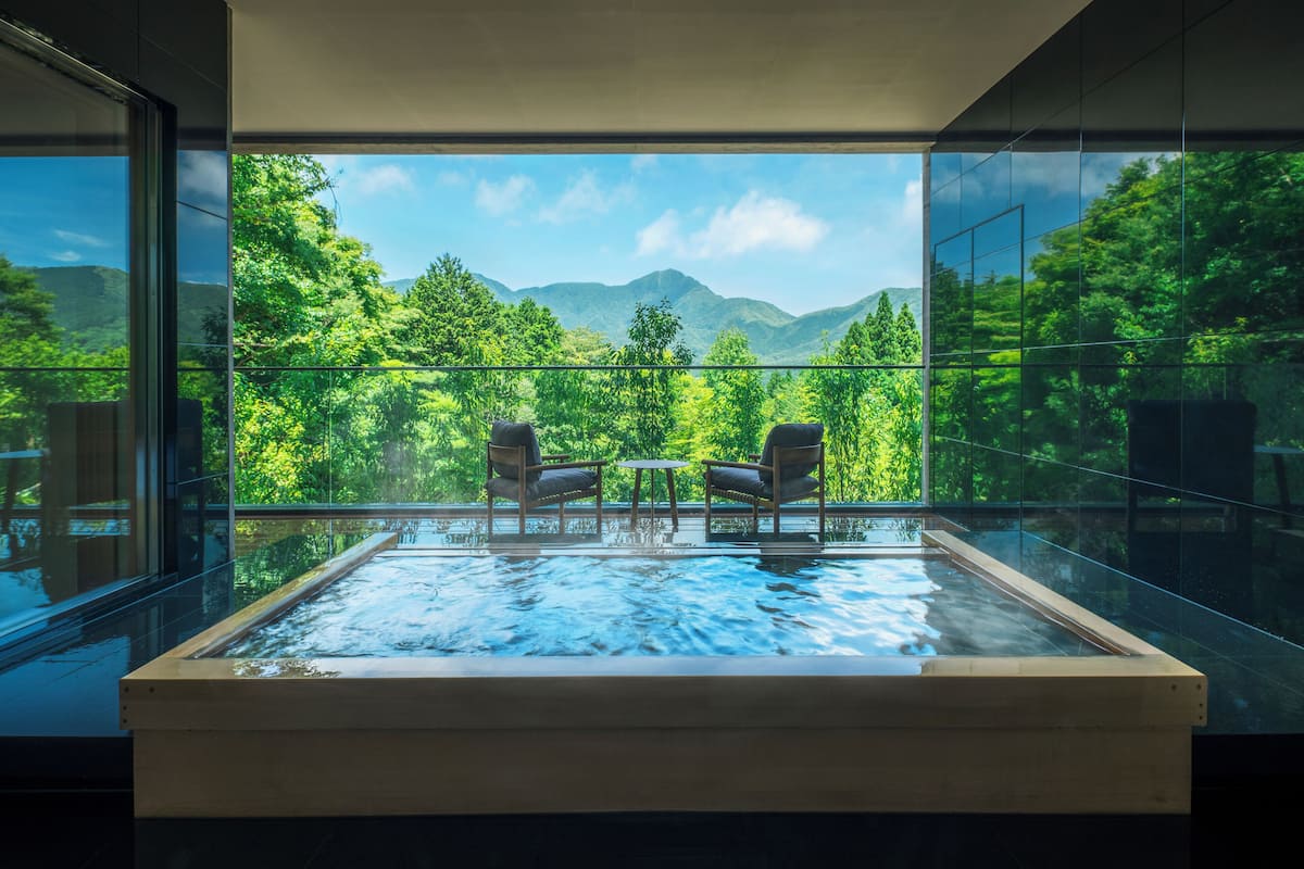 Une piscine intérieure moderne, dotée de deux chaises et d’une table, et entourée de murs de verre qui offrent une vue sur une végétation luxuriante et des montagnes.