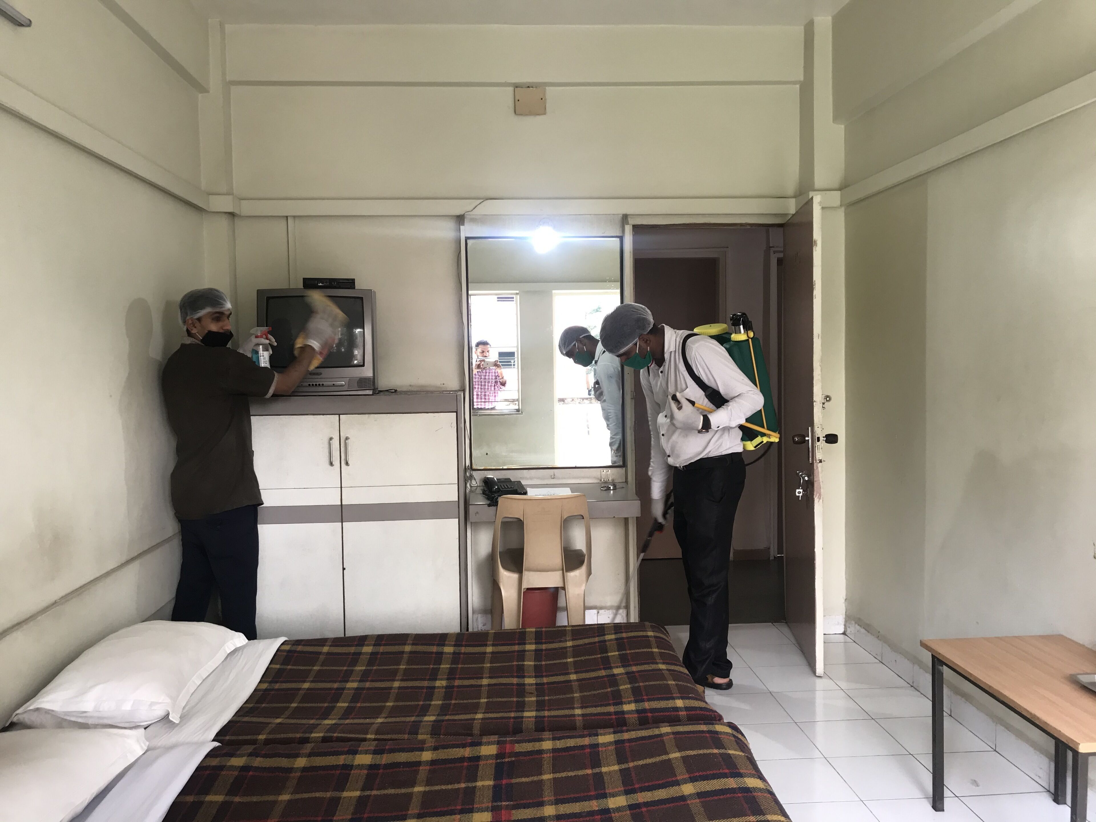 A hotel room with a bed, a television on a cabinet, and a person cleaning with a spray bottle.