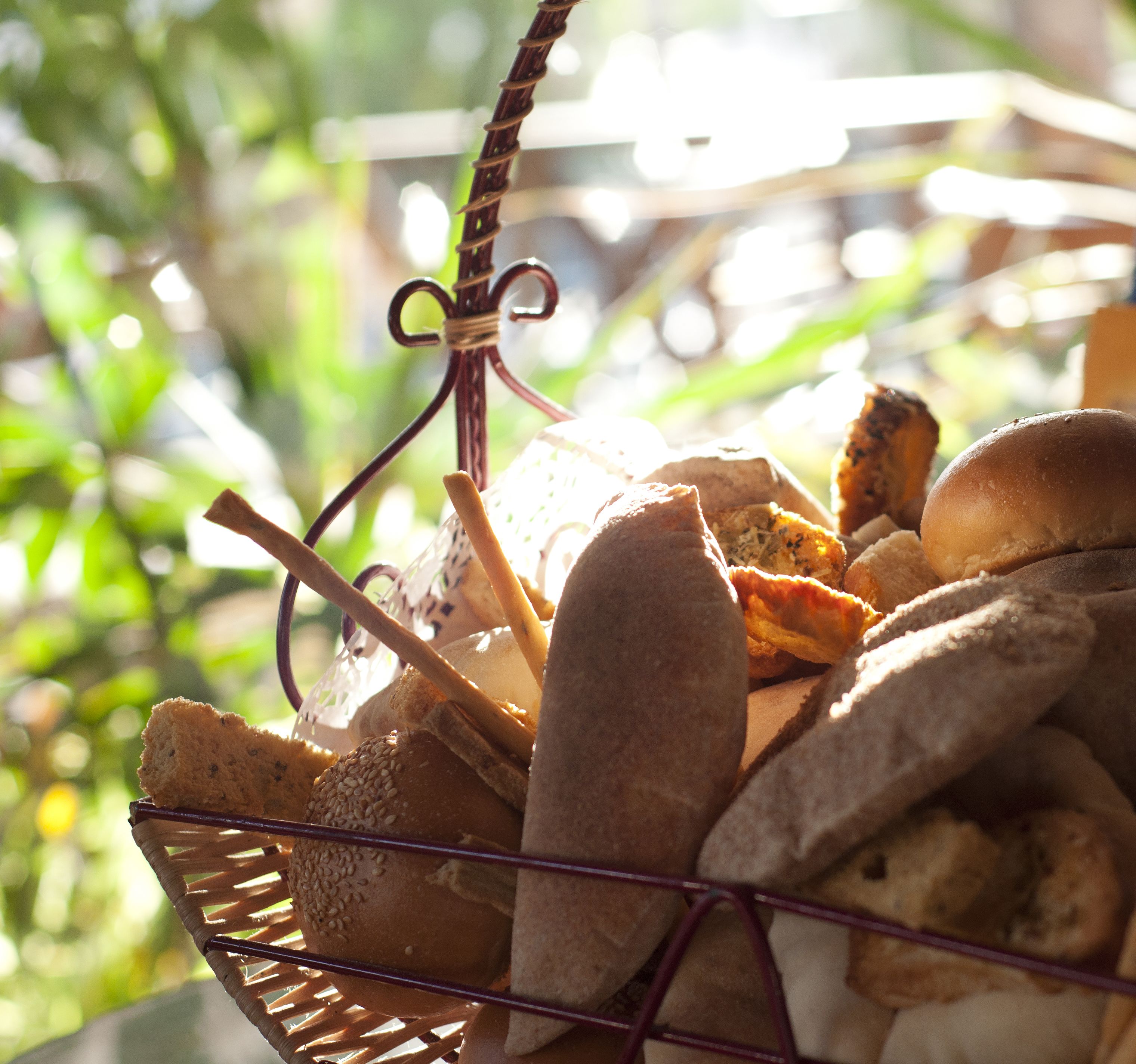 Uma cesta de vime cheia de pães e doces variados, pendurada ao ar livre, com vegetação ao fundo.