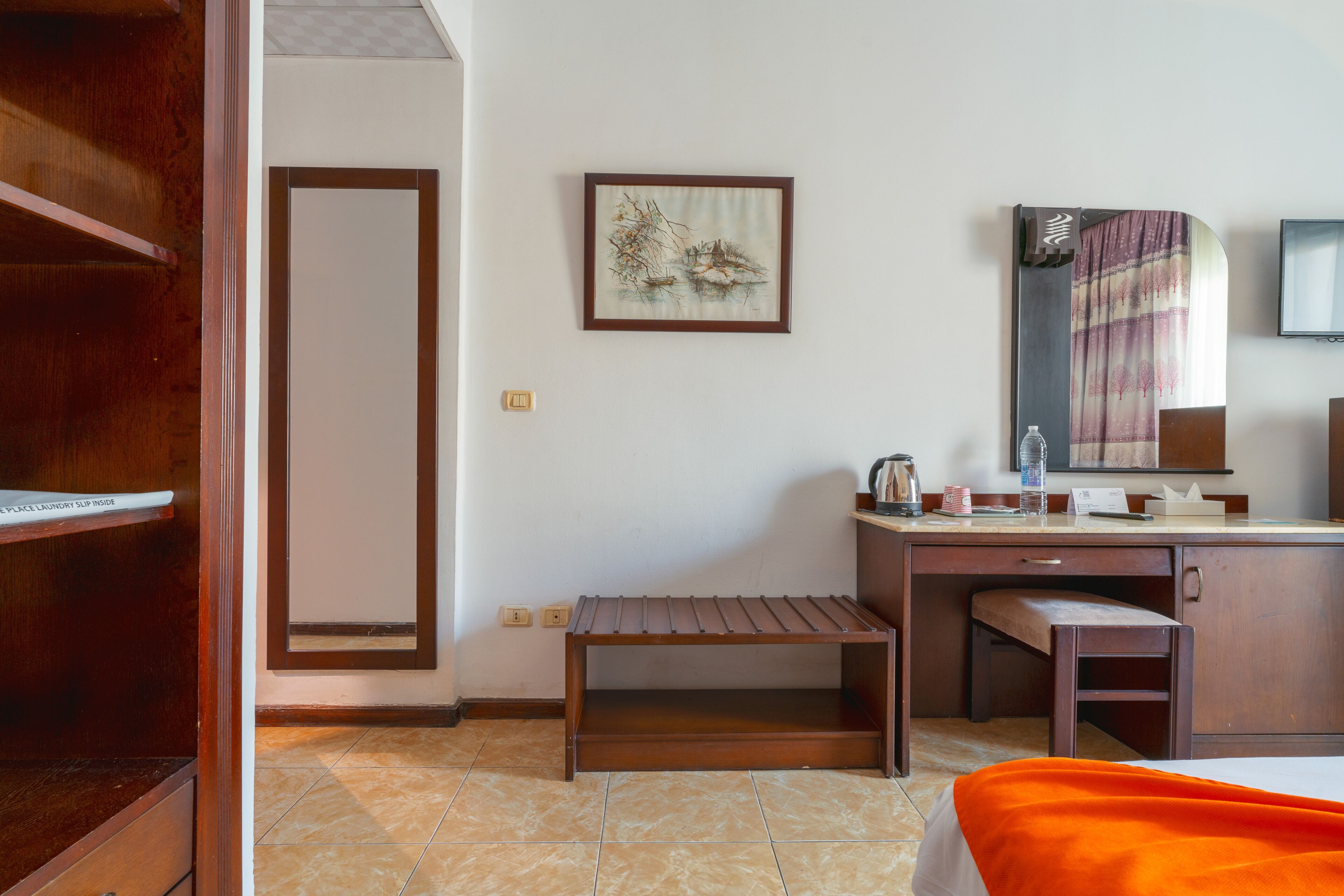 A hotel room with a wooden desk, a bench, a bed with an orange blanket, a framed artwork on the wall, and a television.