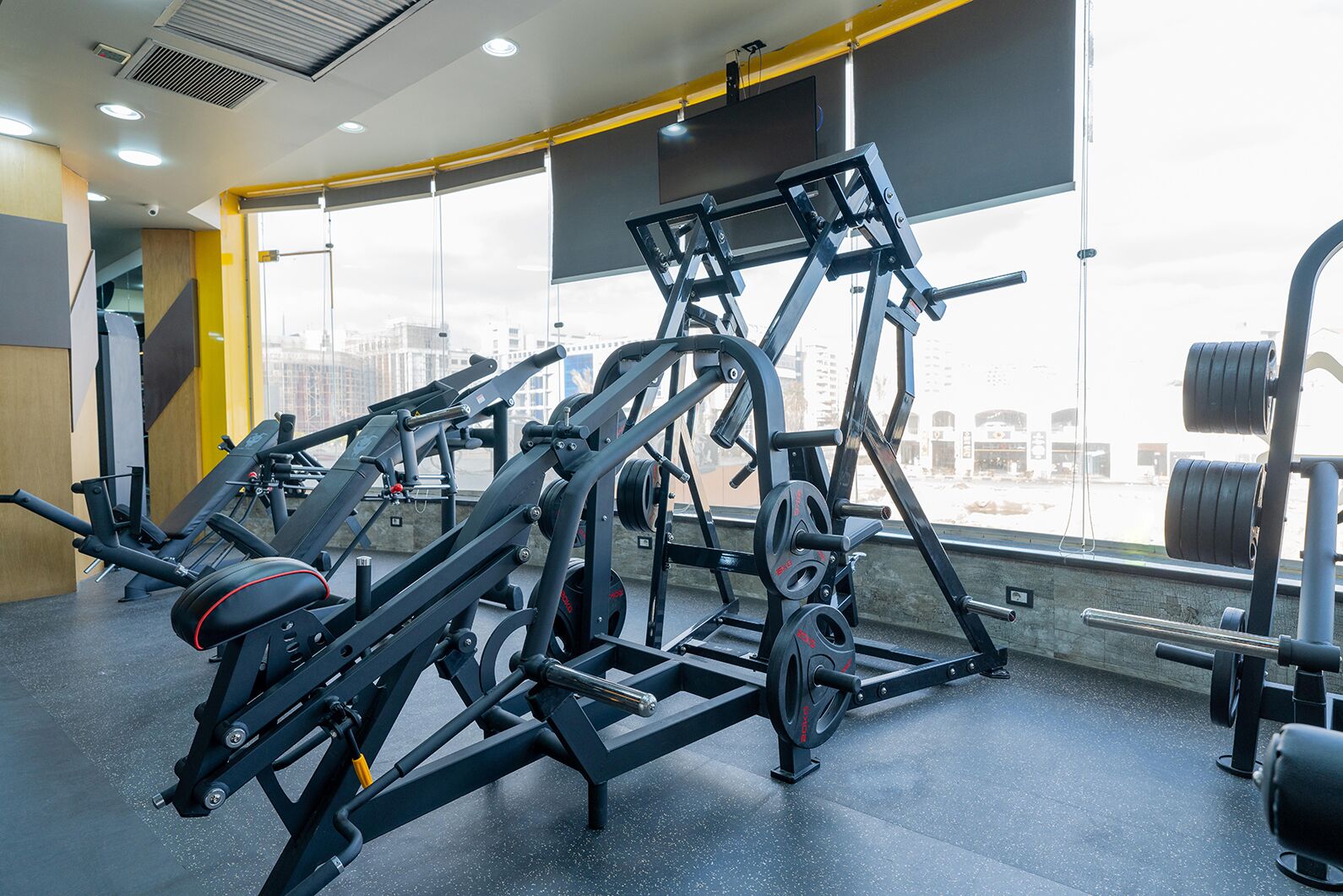Un gimnasio con diversas máquinas de ejercicio, amplias ventanas y vistas a la ciudad.