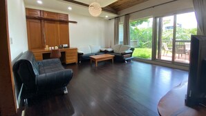 A spacious living area with a wooden ceiling, a large window with a view of greenery, a sofa, a coffee table, and a television.