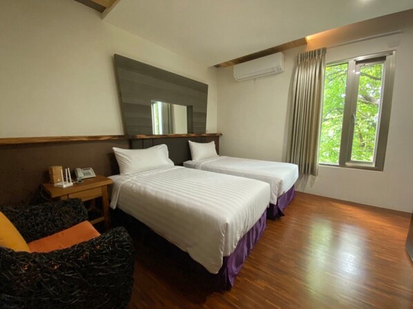 A hotel room with two beds, a wooden headboard, a bedside table with a phone, a mirror, an air conditioning unit, and a window with curtains.