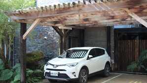 A white SUV parked under a wooden pergola with a stone wall and greenery in the background.