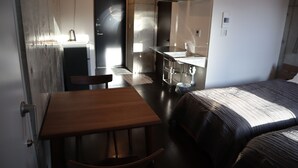 A compact hotel room with a wooden table and chair, a bed with a dark patterned bedspread, a small kitchenette with a sink, and a door leading to a bathroom. - Treeside Hotel & Apartment (Yugawara)