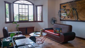 Una sala de estar con un sofá, una mesa con papeles, un ventilador y una bicicleta colgada en la pared.